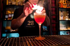 Medium close‑up, eye‑level shot of a bartender torching an orange peel over a red cocktail at 28 HongKong Street, Singapore, with illuminated spirit shelves in the background.