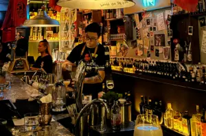 Eye-level medium shot of a bartender crafting cocktails behind a marble bar counter, surrounded by backlit spirits and eclectic décor at a trendy Singapore cocktail bar.