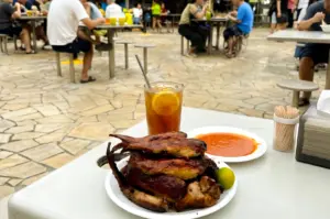 Eye‑level close‑up shot of a hawker center meal featuring roasted or braised meat served on a white plate with chili sauce, calamansi, and a glass of iced lemon tea, set against a busy food court background with communal seating.