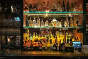 Straight-on close-up shot of a well-stocked back bar in Singapore, featuring rows of premium liquor bottles and glassware under warm accent lighting.