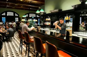 Wide-angle, eye-level interior shot of a classic cocktail bar with patterned tile floors, a bartender shaking drinks behind the counter, and patrons enjoying a relaxed evening atmosphere.