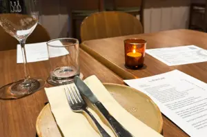 Eye-level close-up shot of a warmly lit restaurant table set with a ceramic plate, neatly placed cutlery on a napkin, wine glass, water glass, candlelight, and printed menu, creating an intimate fine‑dining atmosphere.