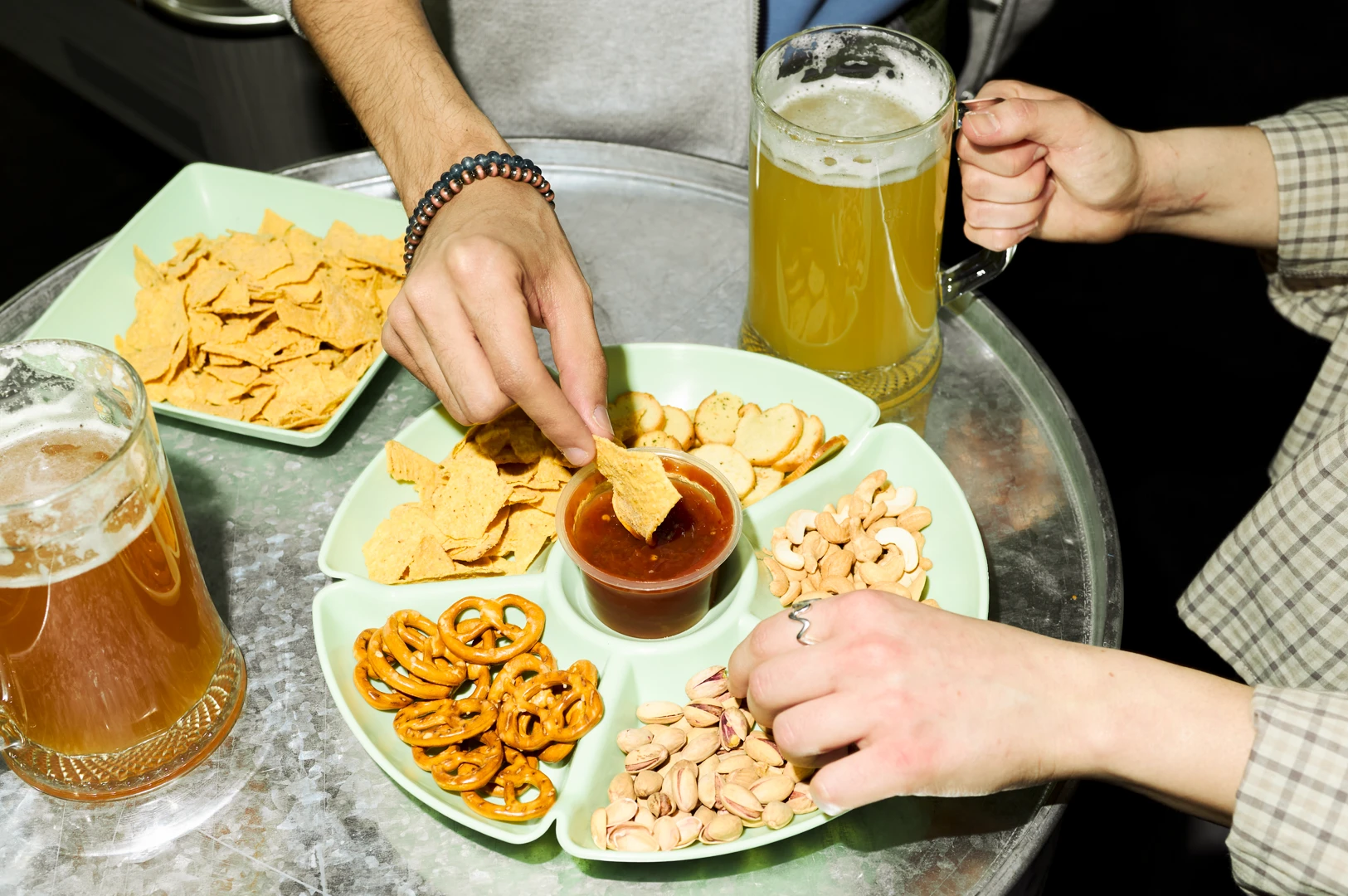Top-down, slightly angled view of hands sharing bar snacks including tortilla chips, pretzels, pistachios, and dipping sauce on a small table with frosty mugs of beer, highlighting casual bar food perfect for drinking.