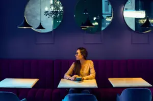 Wide frontal shot of a person sitting alone at a café booth with a cup on the table, surrounded by purple upholstered seating, marble tables, hanging pendant lights, and circular wall mirrors in a dimly lit interior.