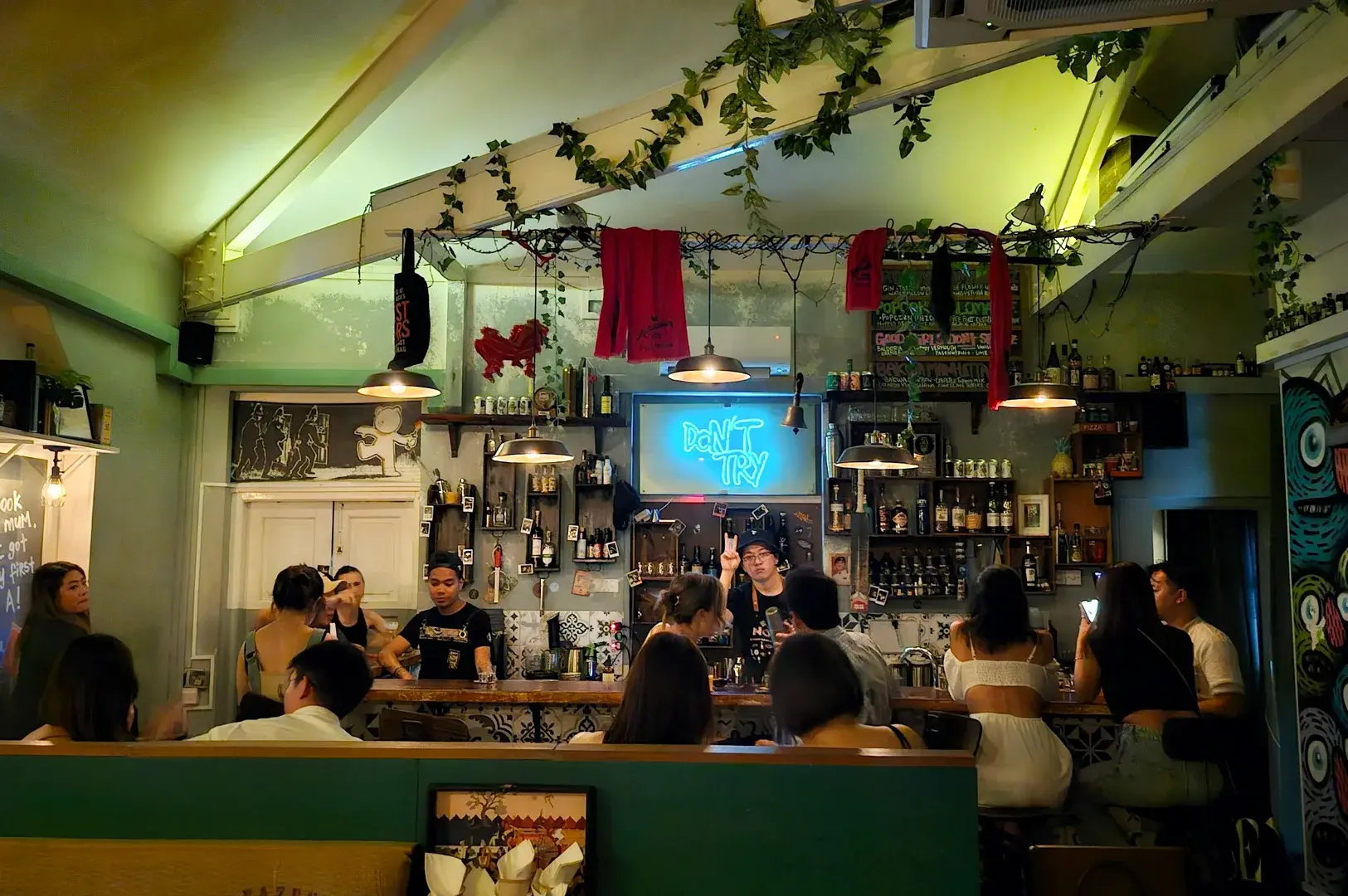 Eye-level wide shot of a crowded casual bar with patrons gathered around a central counter under warm lighting and decorative greenery.