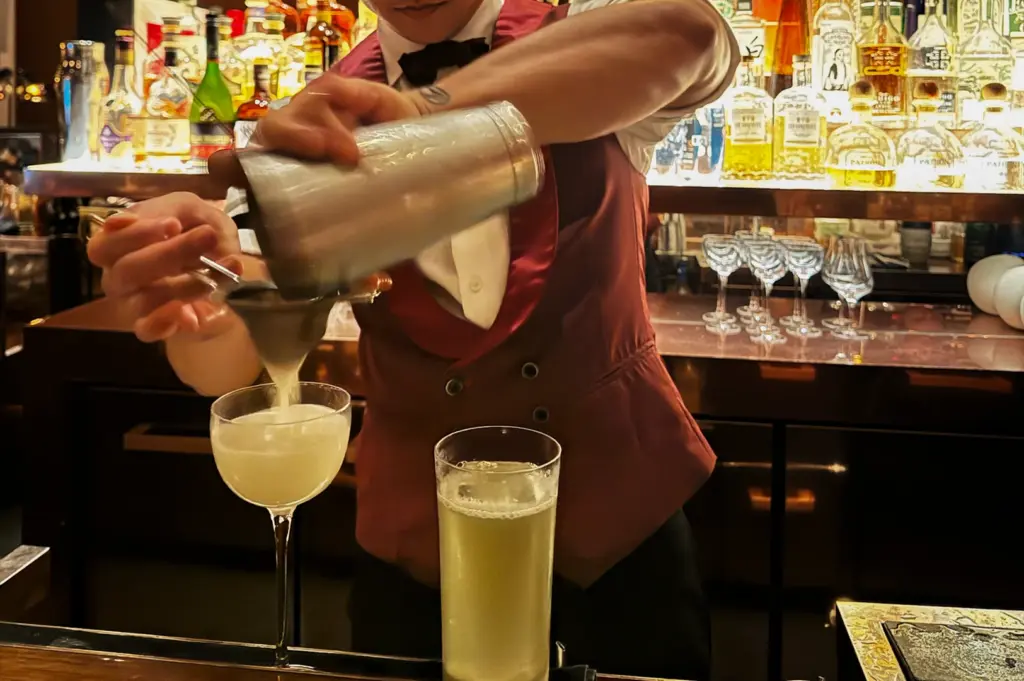 Medium close-up, eye-level shot of a bartender shaking and pouring cocktails at a Singapore bar, with illuminated spirit bottles lining the back bar.