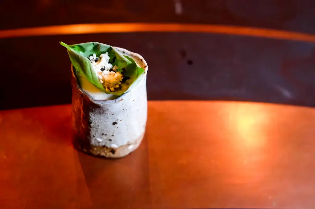 Close-up, eye-level shot of a minimalist cocktail served in a ceramic cup, garnished with ants, fresh green leaves, and foam, presented on a warm copper bar top in a modern craft cocktail bar.