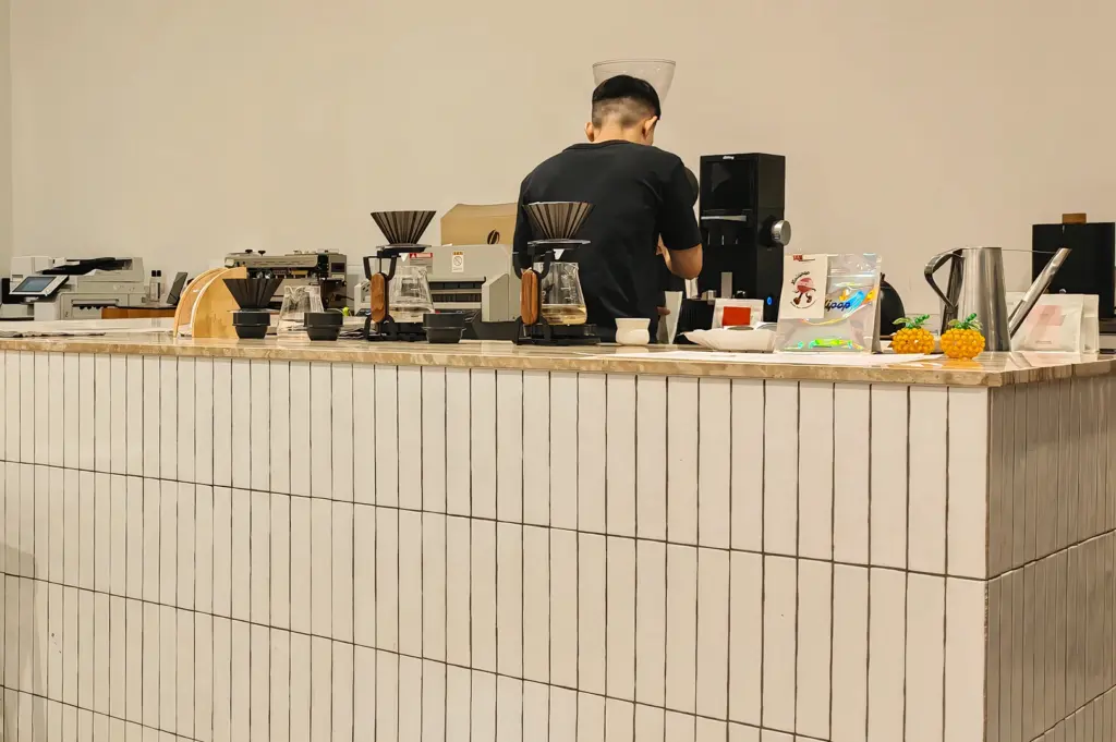 Wide-angle interior shot of a minimalist coffee bar with a tiled counter, pour-over coffee equipment, espresso machines, and a barista working behind the counter in a modern café setting.