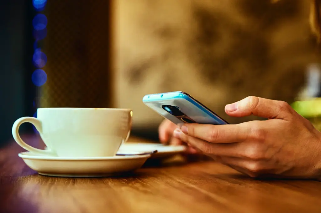 Close-up side-angle shot of a hand holding a smartphone next to a white coffee cup and saucer on a wooden café table with warm lighting and blurred background.