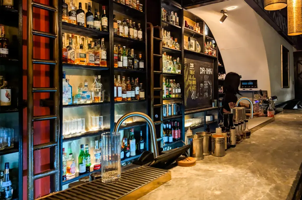 Wide‑angle, eye‑level view of the bar counter at 28 HongKong Street, showcasing backlit shelves of premium spirits, industrial shelving, and an intimate cocktail‑bar setting.