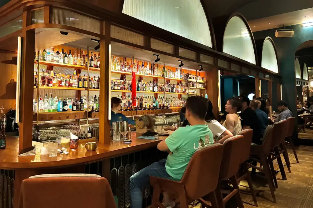 Wide-angle, eye-level shot of a bustling Singapore cocktail bar with guests seated along a wooden counter, backlit shelves of spirits, and warm ambient lighting.