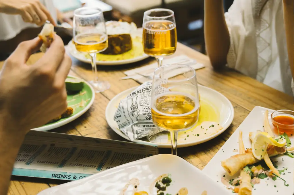 Over-the-table eye-level view of friends sharing small plates and appetizers with glasses of white wine at a wooden bar table, capturing a relaxed social dining experience.
