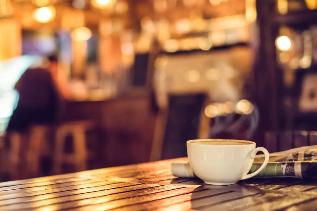 Table-level close-up shot of a white coffee cup beside a folded newspaper on a wooden café table at night, with warm ambient lighting and a softly blurred café interior in the background.