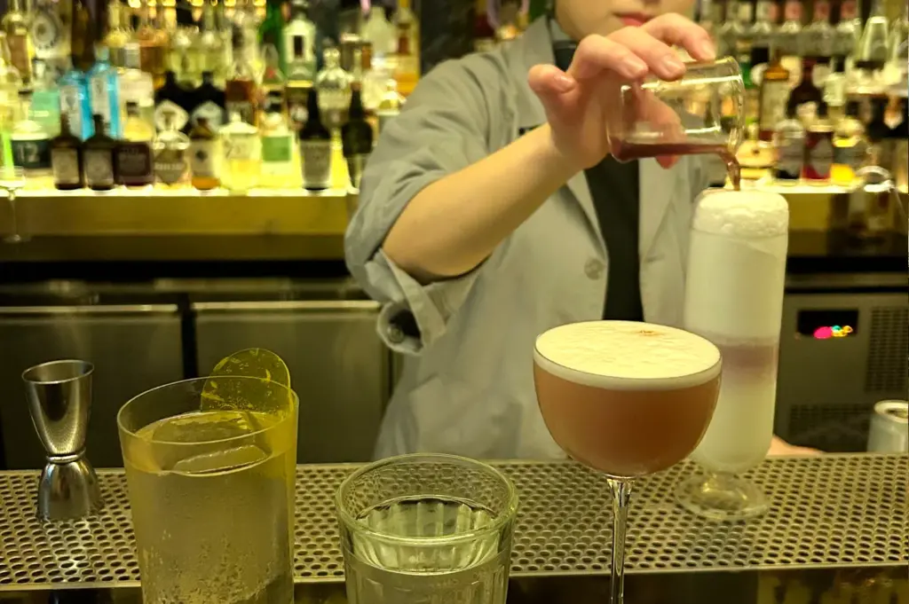 Medium close-up, eye-level shot of a bartender carefully pouring an ingredient into a foamy cocktail, with multiple finished drinks lined up on the bar counter and backlit spirits behind.