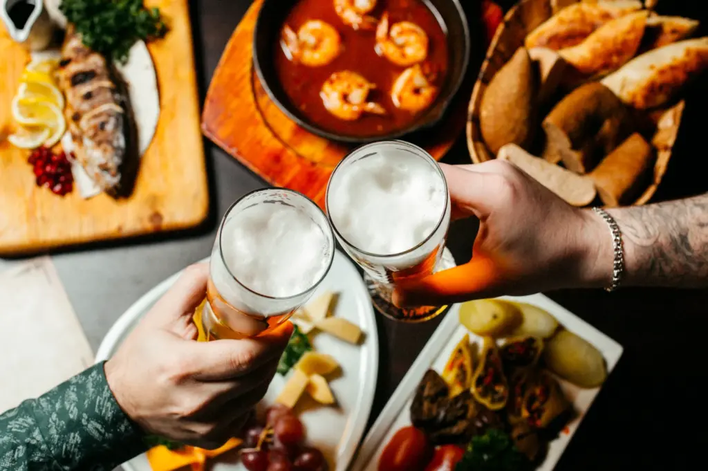 Top-down overhead view of two people clinking glasses of beer over a table filled with shared bar food, including fried snacks, seafood dishes, and small plates, highlighting social bar dining.