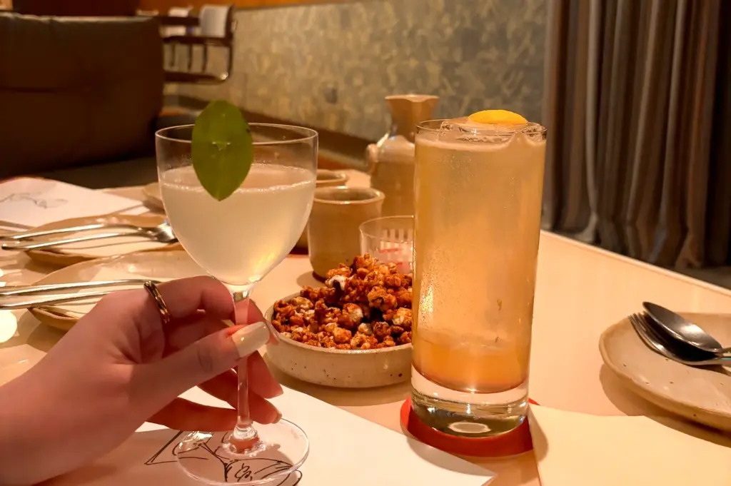 Close-up, eye-level shot of two elegant cocktails on a dining table in a Singapore bar, one held by a guest, accompanied by small bar snacks and minimalist tableware.