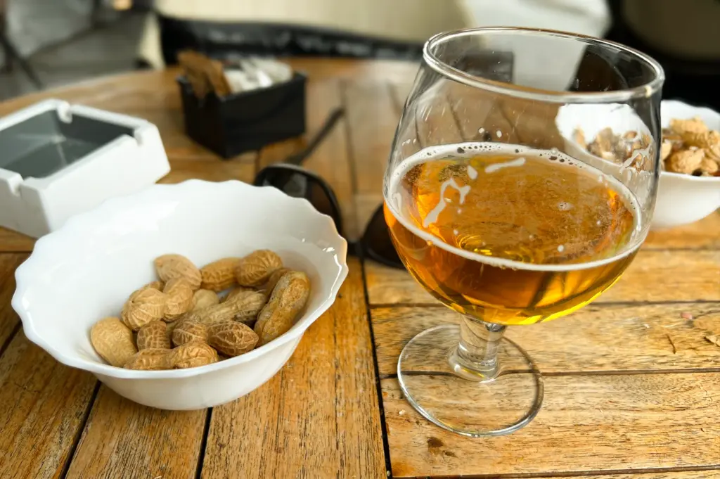 Close-up three-quarter angle of a glass of beer served with a bowl of peanuts on a wooden table, representing simple classic bar snacks that pair well with drinks.