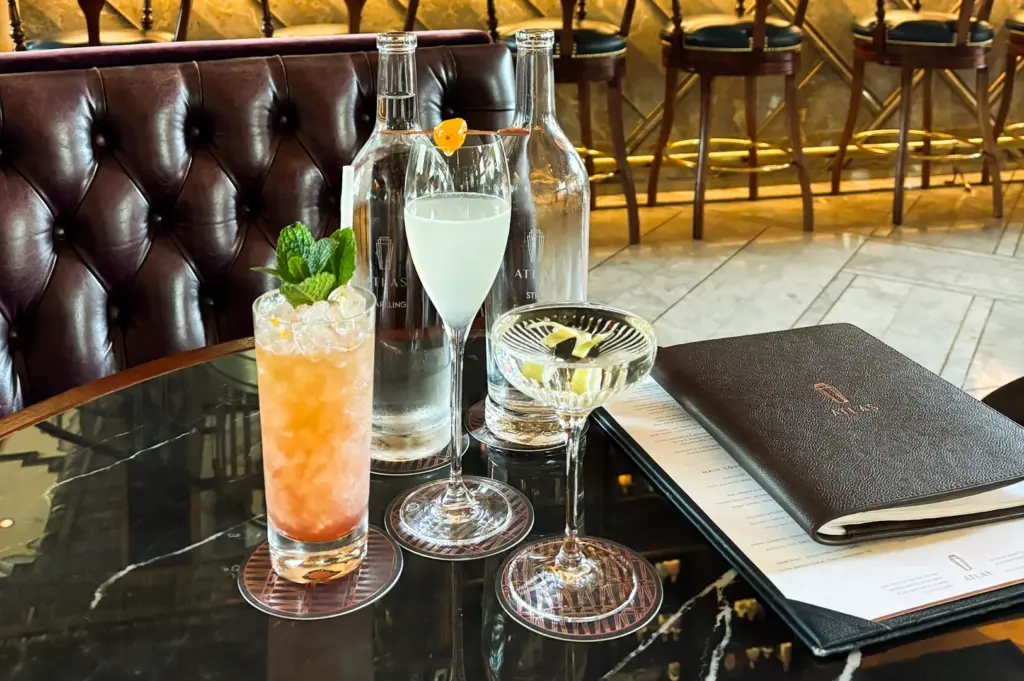 Eye-level medium shot of three signature cocktails displayed on a marble table at an elegant Singapore bar, with leather seating, bottled spirits, and a cocktail menu in the background.