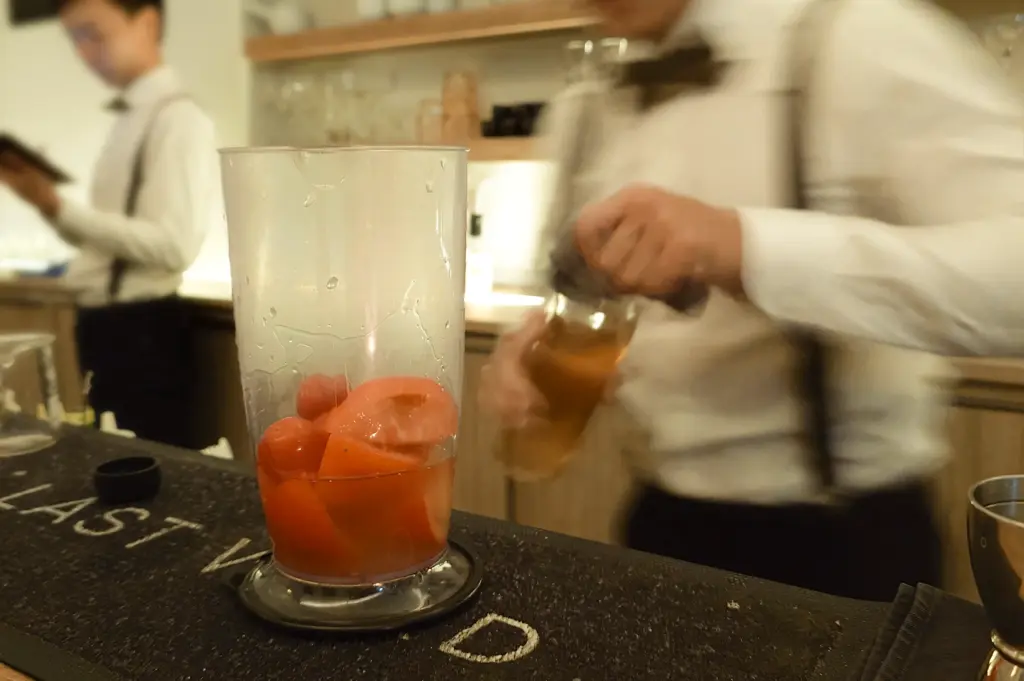 Close-up shot of a bartender preparing a cocktail with grapefruit slices in a mixing glass on a bar counter, with blurred bartenders working in the background, capturing an active bar atmosphere.