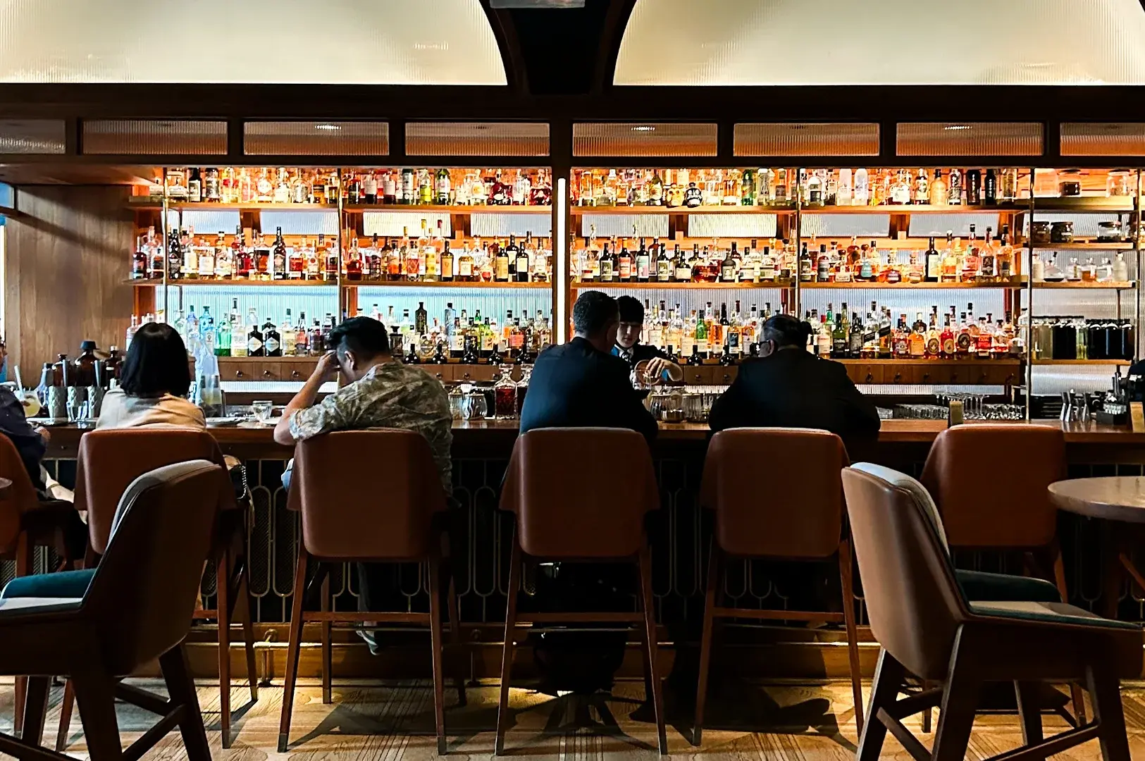 Wide-angle frontal shot of a modern bar interior with multiple patrons seated on leather stools facing a fully stocked backlit liquor shelf.