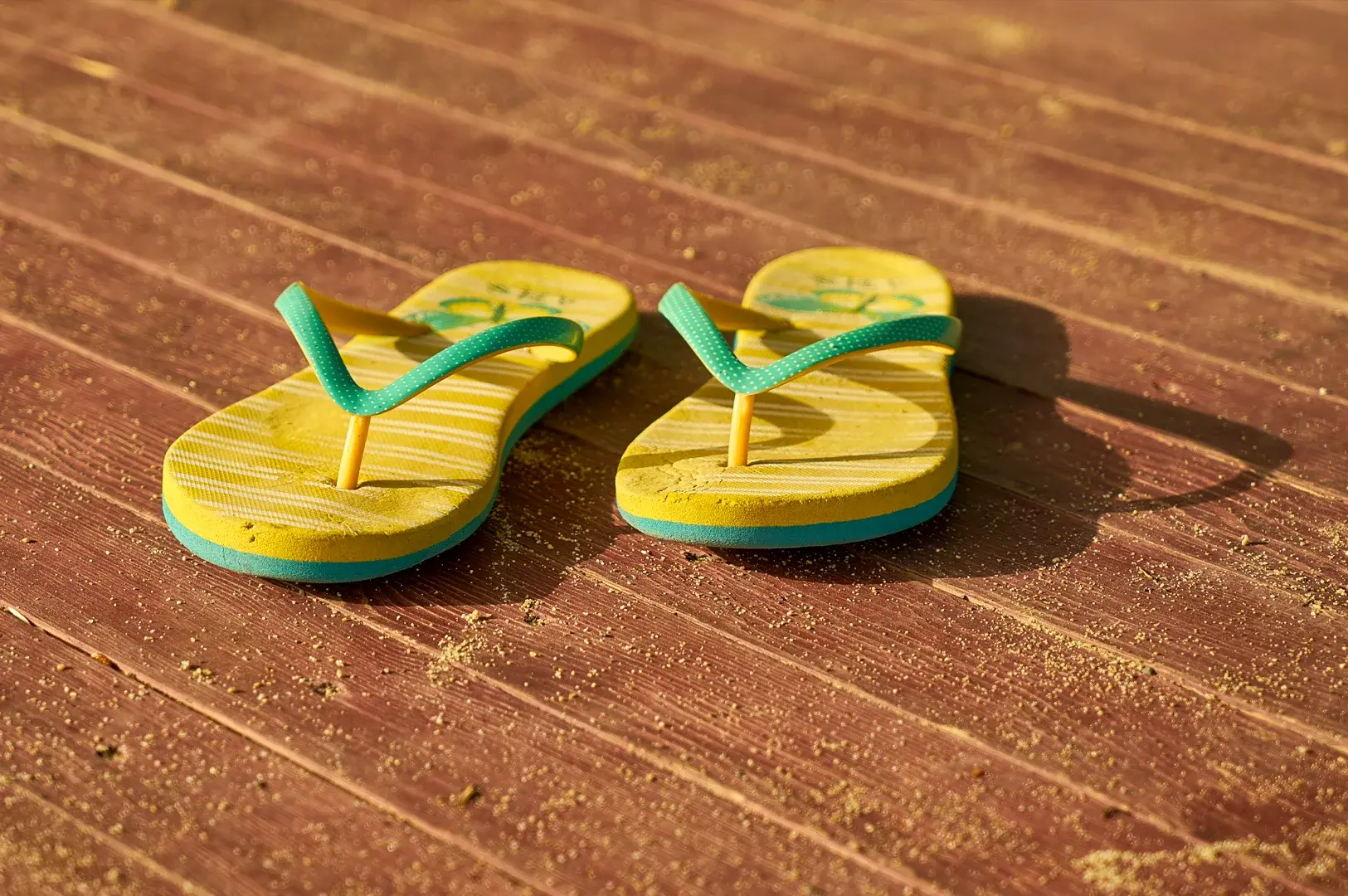 Low-angle close-up shot of yellow and green flip-flops placed on a sunlit wooden floor with visible texture and shadows.