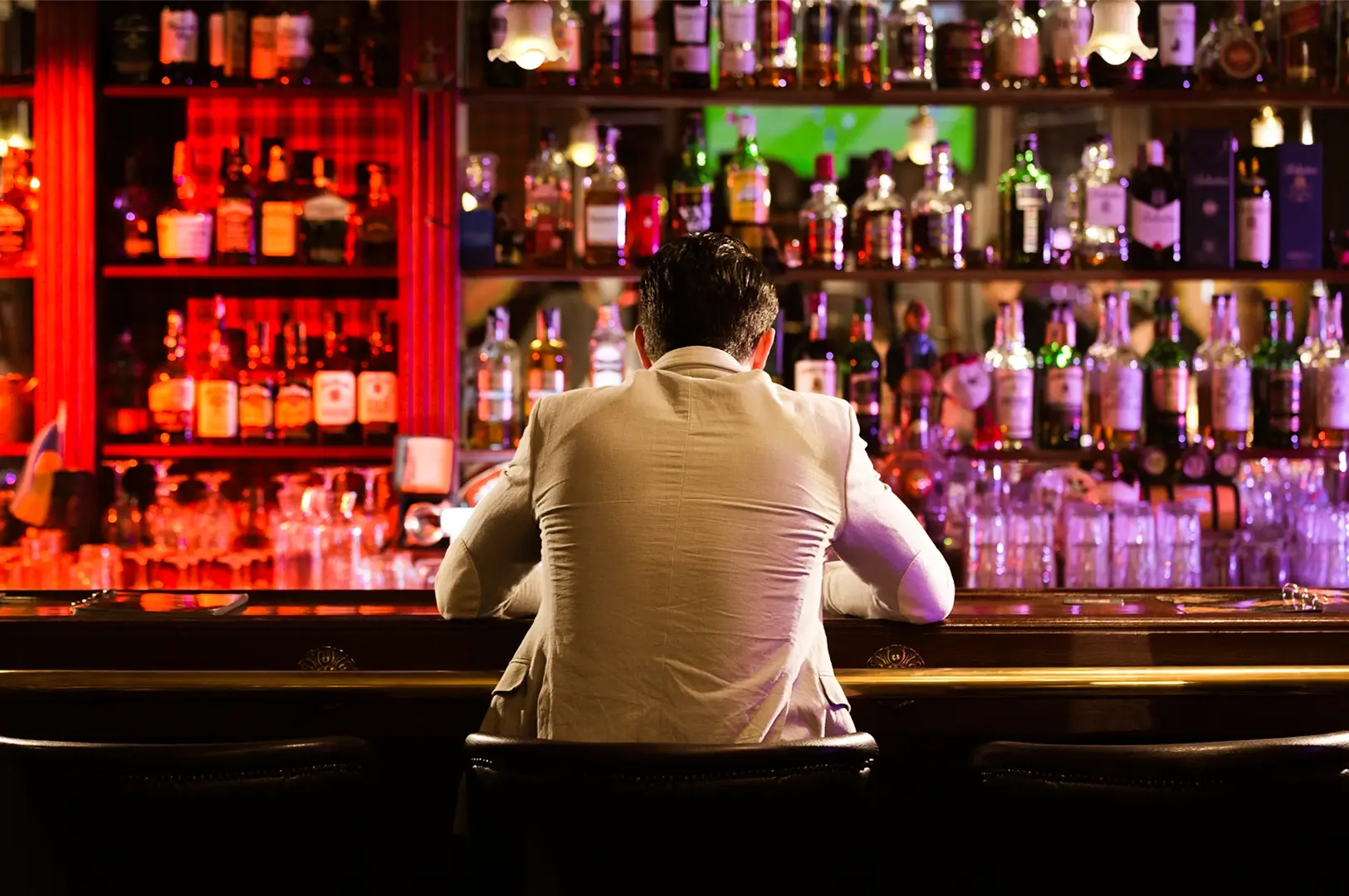 Rear-view medium shot of a person seated at a dimly lit bar counter with rows of liquor bottles illuminated behind the bar.