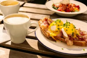 Low-angle close-up of eggs Benedict with poached eggs, crispy bacon, and hollandaise sauce on toasted bread, served with two cups of coffee on a sunlit wooden café table.