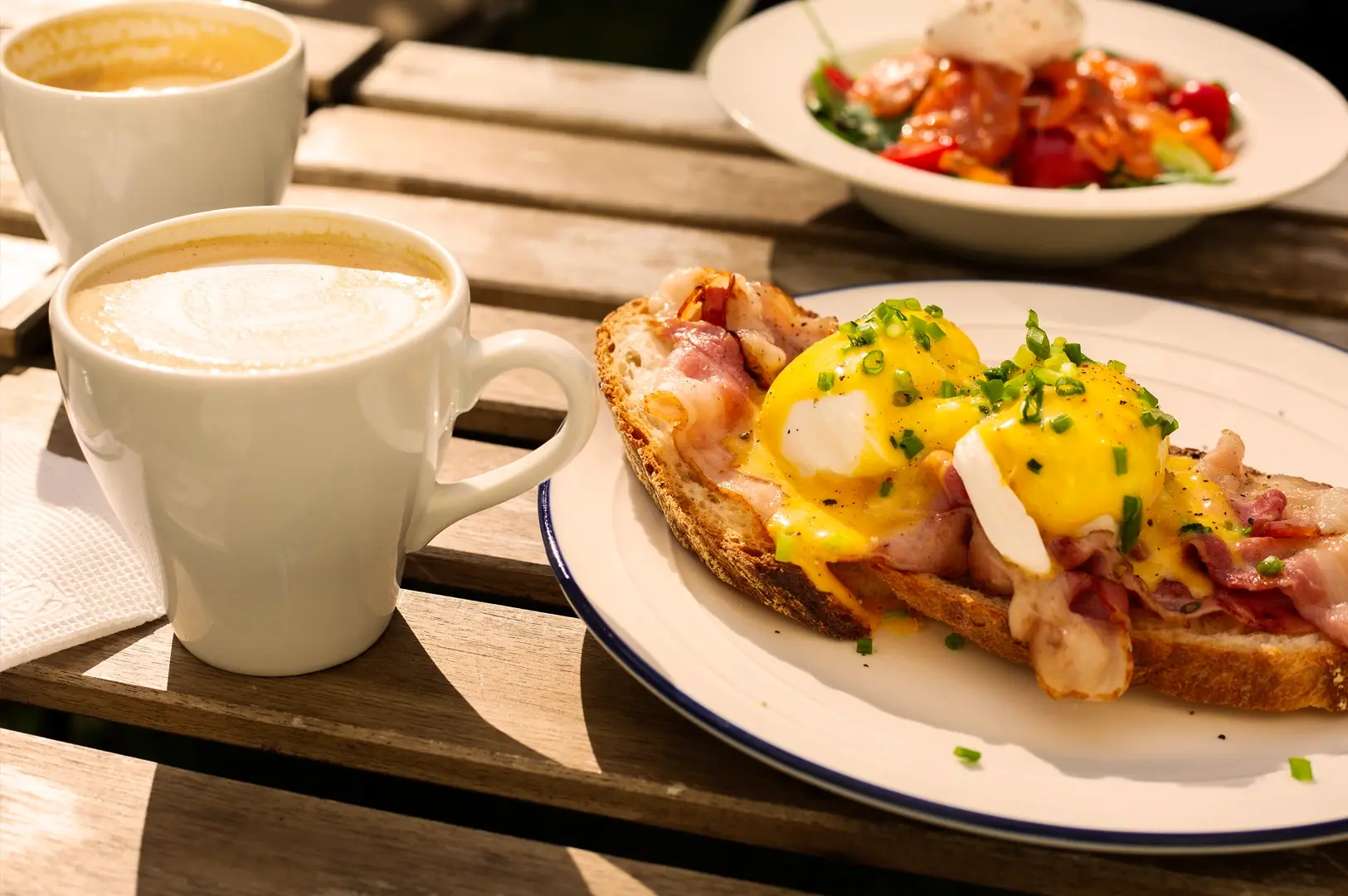 Low-angle close-up of eggs Benedict with poached eggs, crispy bacon, and hollandaise sauce on toasted bread, served with two cups of coffee on a sunlit wooden café table.