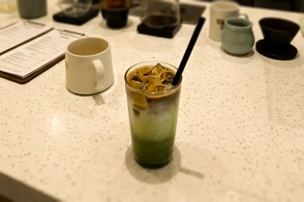 Eye-level close-up of a layered iced matcha espresso latte with ice and a straw on a white speckled café table, with coffee drippers and ceramic mugs softly blurred in the background.