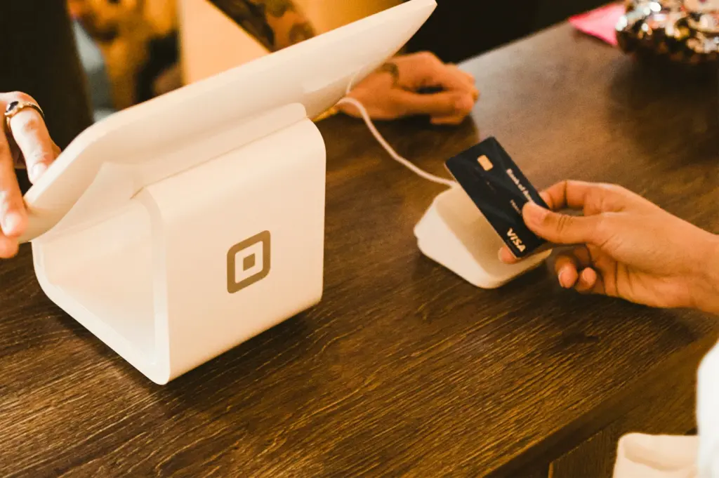 Close-up, eye-level angle of a hand tapping a credit card on a contactless payment terminal beside a Square POS system in a café, illustrating paying for meals and authentic dining experience.