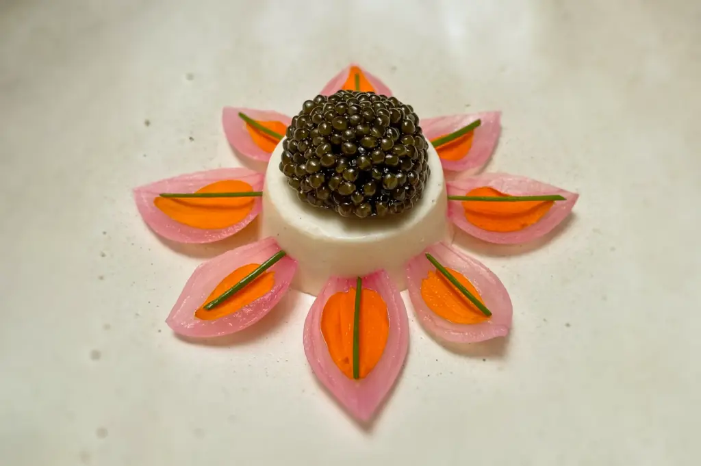 Overhead (top-down) shot of an elegant fine dining caviar appetizer, featuring a creamy white mousse topped with glossy black caviar, surrounded by symmetrical pickled pink petals, carrot slices, and chive garnish arranged in a floral design.