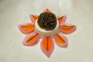 Overhead (top-down) shot of an elegant fine dining caviar appetizer, featuring a creamy white mousse topped with glossy black caviar, surrounded by symmetrical pickled pink petals, carrot slices, and chive garnish arranged in a floral design.