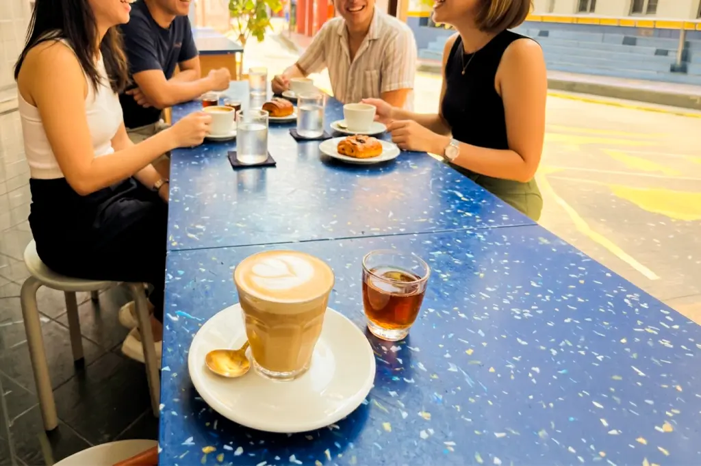 Low-angle foreground shot of latte with latte art on café table, with group of people chatting and pastries in background, highlighting social dining and food influencer photography lifestyle