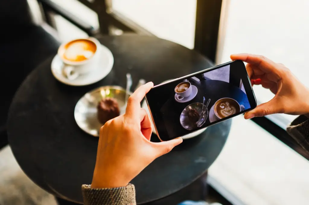 Overhead close-up shot of hands holding a smartphone and photographing cups of coffee and a dessert on a round café table near a window.