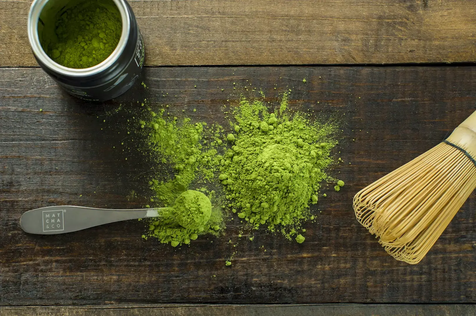 Top-down flat lay of vibrant green matcha powder with scoop, bamboo whisk, and container on rustic wooden surface, representing high-quality matcha powder for lattes and tea