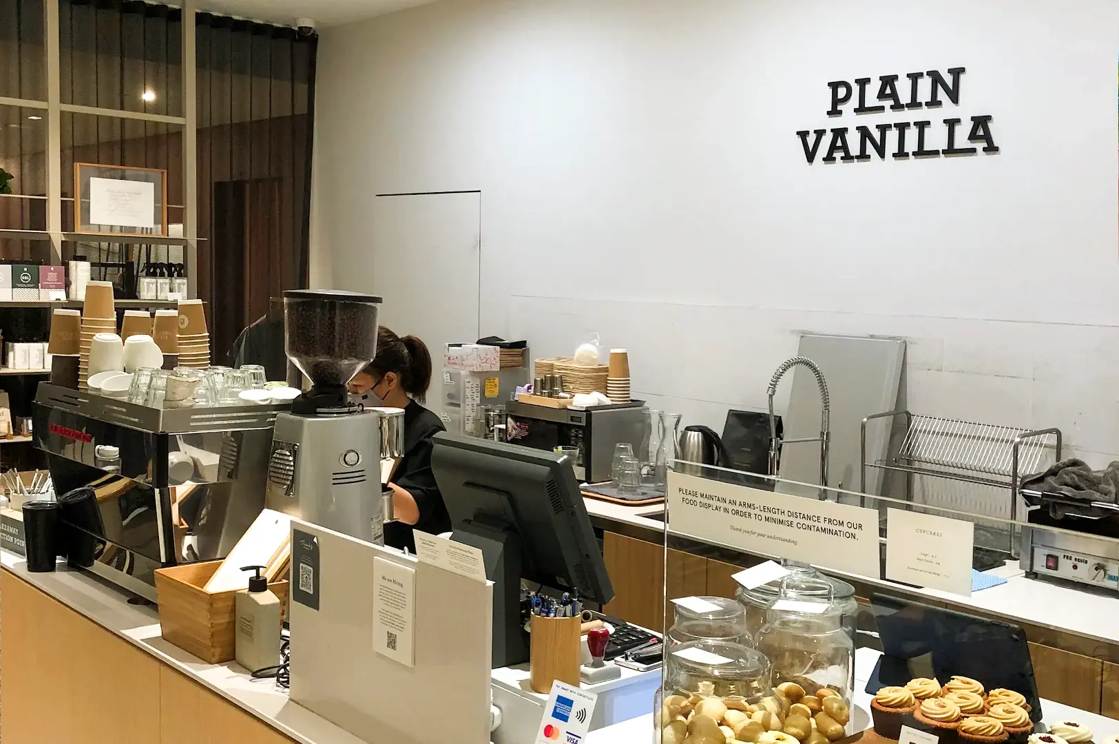 Wide-angle eye-level shot of Plain Vanilla Bakery counter in Singapore, minimalist cafe interior with barista station, pastries, coffee equipment, and cozy Orchard Road brunch atmosphere