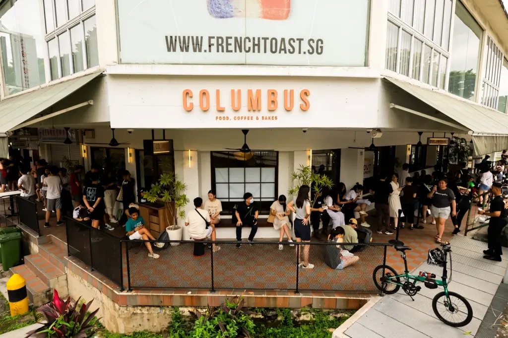 Wide-angle eye-level shot of crowded Columbus Coffee Co in Upper Thomson Singapore, showing long weekend brunch queue and outdoor seating filled with customers, highlighting popular café culture and busy brunch scene