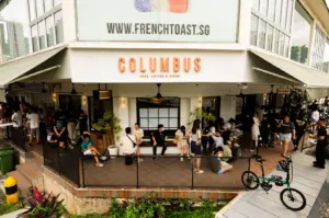 Wide-angle eye-level shot of crowded Columbus Coffee Co in Upper Thomson Singapore, showing long weekend brunch queue and outdoor seating filled with customers, highlighting popular café culture and busy brunch scene