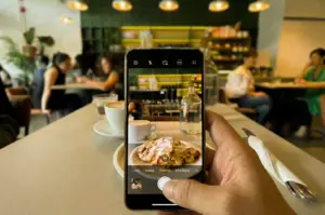 An eye-level, first-person point-of-view (POV) shot focusing on a person's hand holding a smartphone to photograph an almond croissant and coffee at Atlas Coffeehouse in Bukit Timah, with the phone screen perfectly framing the food while the busy cafe background remains softly out of focus.