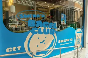 An eye-level, medium exterior shot of the Smear'd Bagel Club storefront window, decorated with vibrant blue-and-white graphics, a cartoon bagel character, a QR menu code, and a metallic handrail in the foreground.