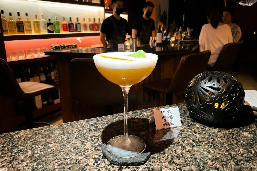 Eye-level medium shot of a frothy craft cocktail in a coupe glass on a polished bar counter, with bartenders and backlit liquor shelves in the background, highlighting modern cocktail bar experience and relaxed premium dining atmosphere