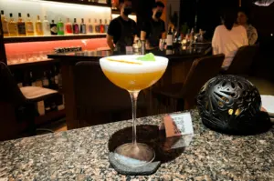 Eye-level medium shot of a frothy craft cocktail in a coupe glass on a polished bar counter, with bartenders and backlit liquor shelves in the background, highlighting modern cocktail bar experience and relaxed premium dining atmosphere