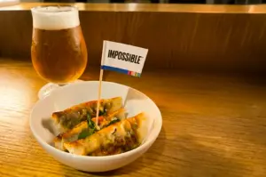 Eye-level close-up shot of spicy Sichuan dumplings in a white bowl with an "Impossible" flag, served at Dumpling Darlings Singapore, paired with a cold draft beer on a wooden counter in a warm, cozy restaurant setting