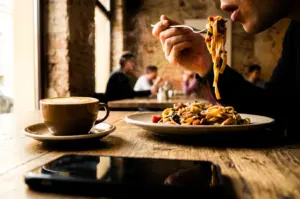 A low-angle, medium close-up shot of a diner eating steaming pasta at a rustic wooden cafe table, capturing noodles lifted on a fork near their mouth, with a ceramic cup of hot latte and a smartphone lying face-down in the blurred foreground.