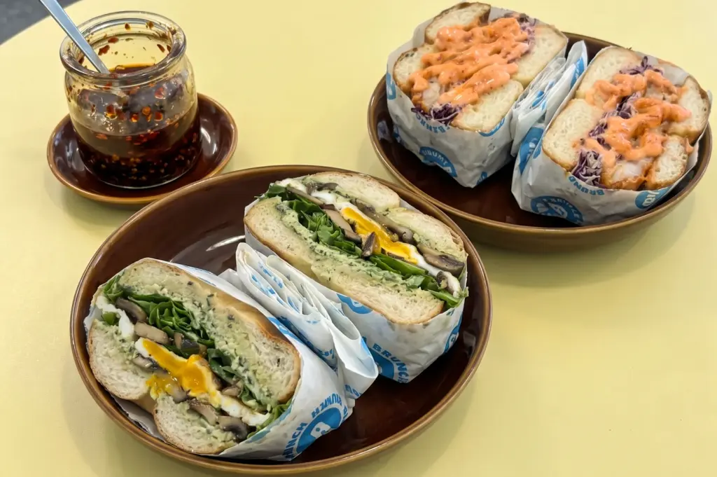 A high-angle, close-up shot of two brown plates on a pale yellow table featuring halved Smear'd savory bagels—one filled with egg, mushrooms, and greens, and the other topped with creamy orange mentaiko sauce—next to a glass jar of chili oil.