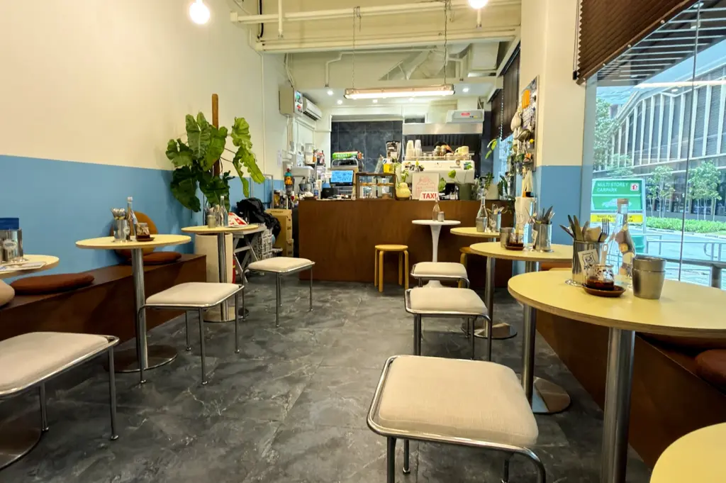 An eye-level, wide-angle interior shot of the compact Smear'd Coffee Bagels café, showcasing minimalist yellow circular tables, small square stools, blue-and-white dual-toned walls, and a wooden ordering counter at the back.