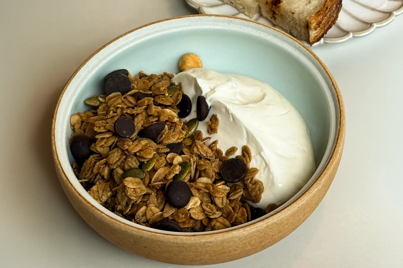A high-angle, close-up shot of an artisanal Dearborn granola bowl, showcasing crunchy dark chocolate hazelnut granola and pumpkin seeds served with a generous scoop of thick, smooth yogurt in a ceramic bowl.