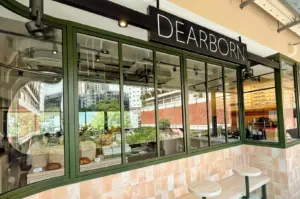 An eye-level, wide-angle exterior shot of Dearborn Breakfast Club's modern facade at New Bahru, highlighting large olive-green framed glass windows, outdoor counter seating, and a prominent black overhead sign with white 'DEARBORN' branding.