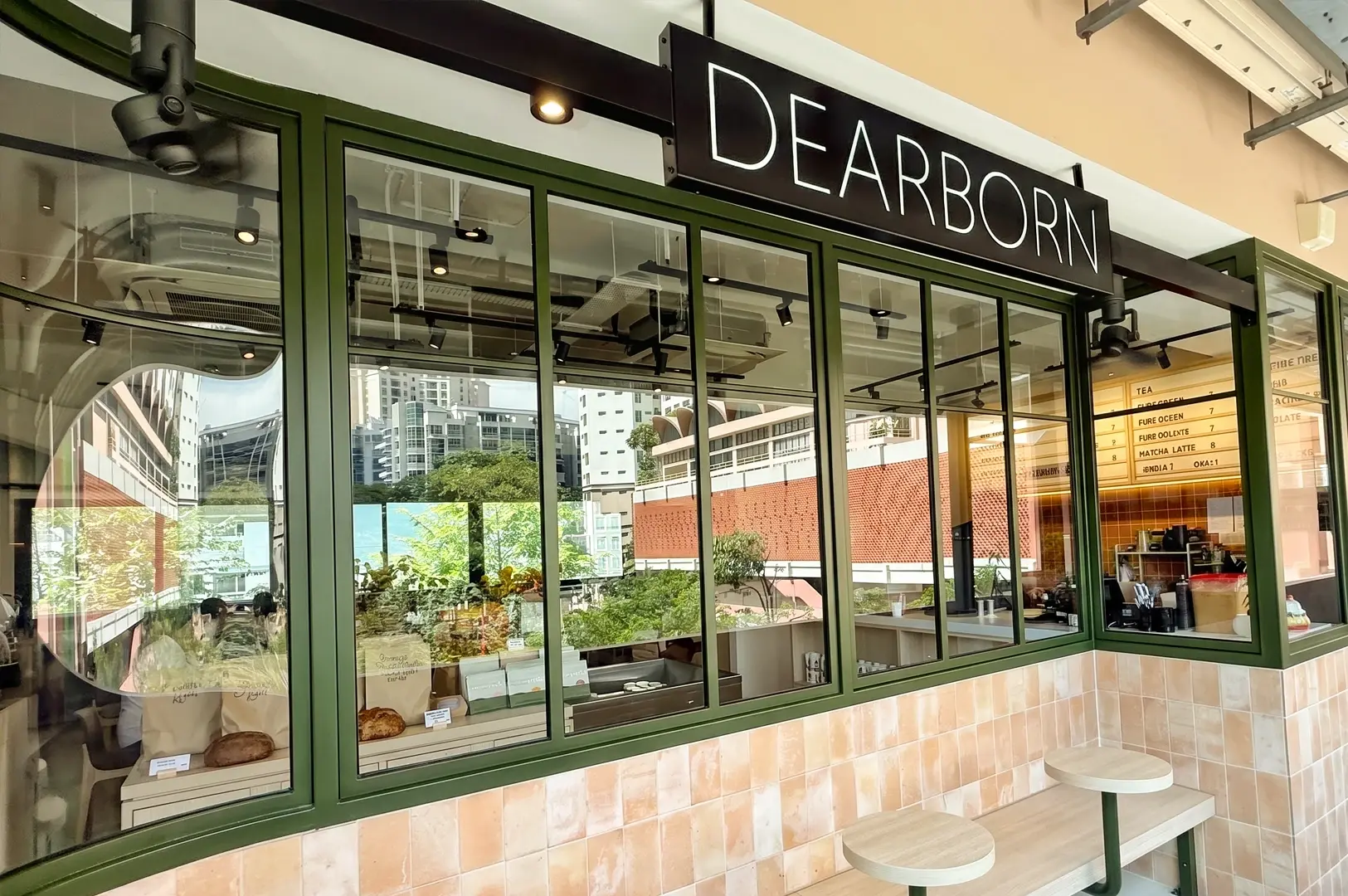 An eye-level, wide-angle exterior shot of Dearborn Breakfast Club's modern facade at New Bahru, highlighting large olive-green framed glass windows, outdoor counter seating, and a prominent black overhead sign with white 'DEARBORN' branding.