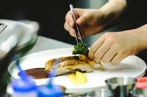 Close-up, eye-level angle of a chef plating a dish, with hands using metal tweezers to place a small green garnish onto a piece of seared meat on a white plate, accompanied by a smooth yellow puree and dark sauce; blurred kitchen elements and a blue flame visible in the foreground, with warm lighting highlighting textures, gloss, and fine dining presentation.